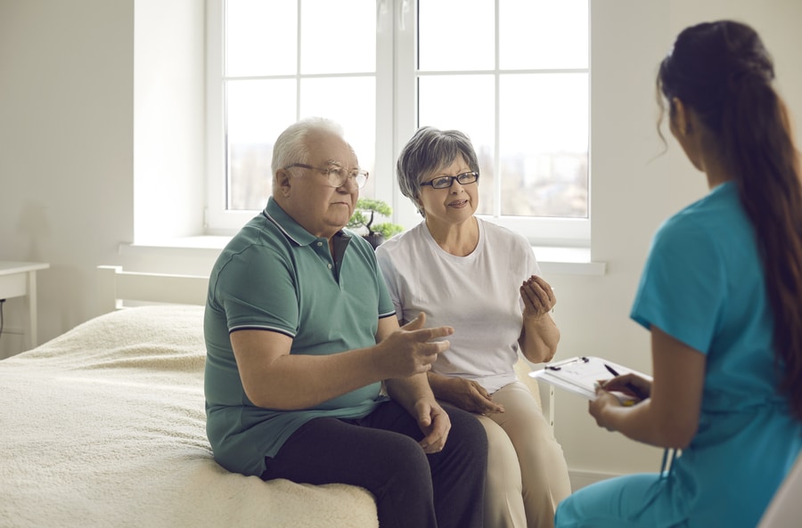 Worried Senior Couple Sitting On Bed And Talking To Nurse Or Fam